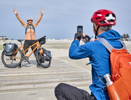 Mann macht von Frau und Rad ein Bild mit dem Wattenmeer im Hintergrund.